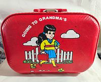 Front view of the red vintage children's suitcase featuring the cartoon girl, white picket fence, flowers, clouds, and 'Going to Grandma's' text.