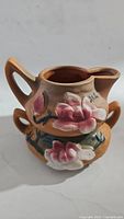 Cream pitcher showing brown glaze with raised pink and white magnolia flowers and green leaves, side view.