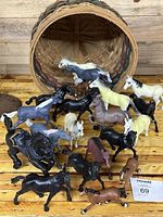 Basket laying on side with 17 vintage horse figurines of various colors and postures displayed around it on a wood surface.