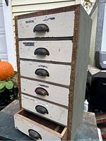 Front angled view of small wooden cabinet showcasing five drawers with black metal cup handles and antique ruler trim around edges. One drawer partially open.