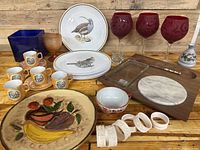 Full lot overview showing bird plates, red wine glasses, blue vase, brown goblet, ceramic fruit plate, cheese board with marble, napkin rings, and decorative tea cups with saucers.