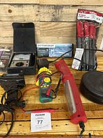 Photo shows a collection of garage tools including a digital caliper, soldering iron, retractable light, a meter with probes, and a black case on a wooden surface.