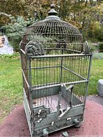 Front perspective showing green chippy wooden cage with decorative metal wire bars, domed top and ornamental elements. Door partially broken.