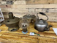 Photo showing antique kettle, brass cooking pots, candle holder, and glass oil lamp on wooden surface with rustic backdrop.