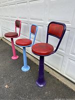 Three vintage Woolworth soda fountain stools arranged outdoors against a white garage door, showing front and side views. Pink, light blue, and dark purple bases. Red vinyl seats and backrests show wear.