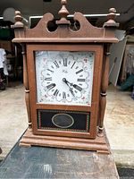 Front view of wooden Welby mantel clock showing decorative top finials, painted clock face with Roman numerals and floral details, wooden base with stepped design