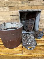 Photo shows a large metal sap bucket, two cast iron pans with one having animal shapes, a weathered wooden crate, and a small metal pulley wheel all arranged against a wooden backdrop.
