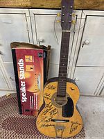 Acoustic guitar leaning against a locker with a box of speaker stands beside it. The guitar has many signatures on the body and a black pickguard.