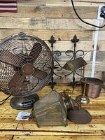 Front view of vintage table fan, brass desk lamp, outdoor light fixture, iron wall hangings, and copper pot arranged on wooden surface