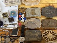 Overview of lot on wooden surface including six metal belt buckles with various western and novelty themes, dice, keychains, wristwatch, and small boxes.