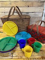 Photo showing the wicker picnic basket, tartan metal basket, colorful divided plastic plates, a blue compartment tray, and plastic cups