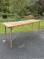 Full view of vintage wallpaper table standing outdoors on pavement showing trapezoidal wooden folding legs and worn wooden top with metal edge on one side.