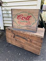 Two wooden crates stacked outdoors on pavement next to a house: a smaller Cott Quality Beverages crate on top and a larger wooden trunk underneath with a faded logo.