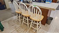 Three wooden bar stools positioned side by side next to a kitchen counter. Backs and legs painted white, seats natural wood.
