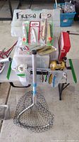 View of fishing lures in original packaging, fishing reel, and landing net on table.
