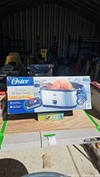 Front view of Oster 20-quart stainless steel roaster oven box, showing product image with cooked chicken inside roaster, and branding.