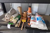Shelf view of painting supplies including brushes, masking tape, rubber tubing, Petrol paint thinner, and plastic drop cloths