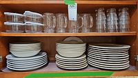 Wooden cabinet shelf showing a collection of white dinner plates, salad plates, bowls, clear glass mugs, and clear plastic tumblers stacked neatly.