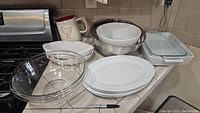 Wide view of large and medium Pyrex glass mixing bowls, white ceramic serving plates/trays, clear rectangular casserole dish with lid, terracotta pitcher with red chicken, and large white mixing bowl with metal rim on kitchen counter.