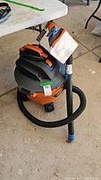 Ridgid black and orange shop vacuum with attached hose and wand on concrete floor near table.