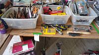 Wide view of three plastic bins containing assorted hand tools including scissors, pliers, wrenches, sockets, and screwdrivers, plus a wire brush and tape measure on sides.