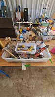 View of the multiple assortments of hand tools including hammers, pliers, staple guns, wrenches, and boxes of staples in plastic bins on a table.