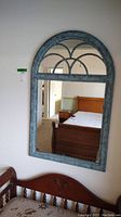 Image of a green framed wall mirror with an arched top and decorative design, mounted on a white wall, reflecting bedroom furniture and window light.
