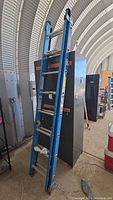 Left side view of the blue Werner fiberglass extension ladder leaning against a machine indoors