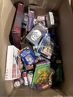 Top view of box containing assorted action figures, collectible cards, card game packs, and cans, with visible Teenage Mutant Ninja Turtles figure and Star Wars figure in packaging.