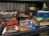 Photo showing stacked board games including Battleship, Trivial Pursuit DVD Star Wars, Lord of the Rings Risk, Scene It?, and a calculator, plus other games on shelf.