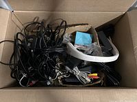 Top view inside a cardboard box showing a chaotic bundle of various black cables, an AC adapter, some wired connectors, and a white plastic item.