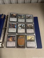 Blue 3-ring binder opened flat displaying plastic sleeves holding 9 Magic: The Gathering cards per page. Cards visible on two pages show various characters, artifacts, and spells.