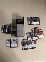 An overhead view showing a large collection of Magic: The Gathering cards spread around a black plastic storage box holding a deck.