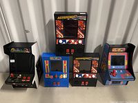 Front view of four small arcade game units, two Asteroids units on right, Ms. Pac-Man in blue, Contra on left.