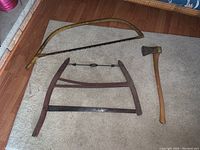 Wide view of the antique bow saw, handsaw, and axe placed on carpeted floor showing overall condition and design.