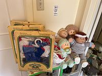 Five Cabbage Patch Kids dolls arranged on a green surface, three are babies with clothes and pacifier, two toddlers with yarn hair and outfits. Several packages of baby clothes are also shown in original boxes.