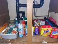 Photo showing cleaning products under kitchen sink including bottle of Cascade dish liquid, Lysol spray, bathroom cleaner, and various sponges and pads.