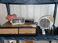 Six items displayed on shelf: piggy bank, Westclox clock, Sylvania clock, electric heater, VHS case with tapes, plastic file holder.