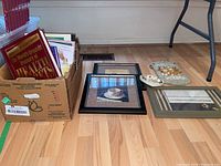 Photo showing a box of cookbooks and Coronation Street magazines on the left and a few framed prints, a wall decor plaque and decorative plate on the floor.