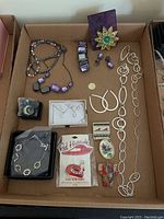 Overview of various costume jewelry pieces laid out in a box, showing necklaces, earrings, a flower brooch, cameo brooch, and Red Hat pin.