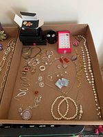 Full overview of assorted costume jewelry in a box including earrings, necklaces, bracelets, brooches, and rings arranged loosely on a flat surface.