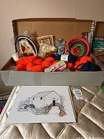 Wide shot of box containing orange and blue yarn skeins, knitting looms, needlework kits, partially finished paintings, and assorted craft supplies.
