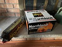 Box of Northland firelogs sitting on a hearth, shows front of box with label and a wrapped bundle leaning against it.