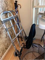 Photo showing collapsible walker with wheels, walking sticks, and mesh basket in corner near window and wallpapered wall.