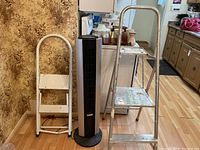 Photo showing the Bionaire tower heater placed between a white two-step ladder and an aluminum ladder, all items visible.