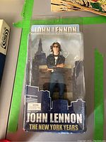 Figure in packaging showing John Lennon wearing a black jacket and sunglasses with New York skyline background.
