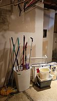 Photo showing mop handles, brooms, and various bins stored on a concrete floor in basement area.