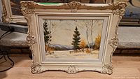 Frontal photo of framed oil painting showing winter landscape scene with pine and birch trees, snow, and mountains under pale sky. Ornate off-white carved wooden frame visible.