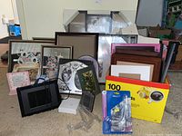 Photo showing assortment of picture frames stacked and leaning against a wall on the floor with a box of photo tabs and some packaging materials.