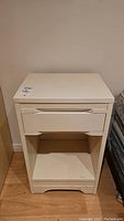 Front view of the first side table showing a painted wood table with a drawer and open shelf. Light cream color, slight wear visible.